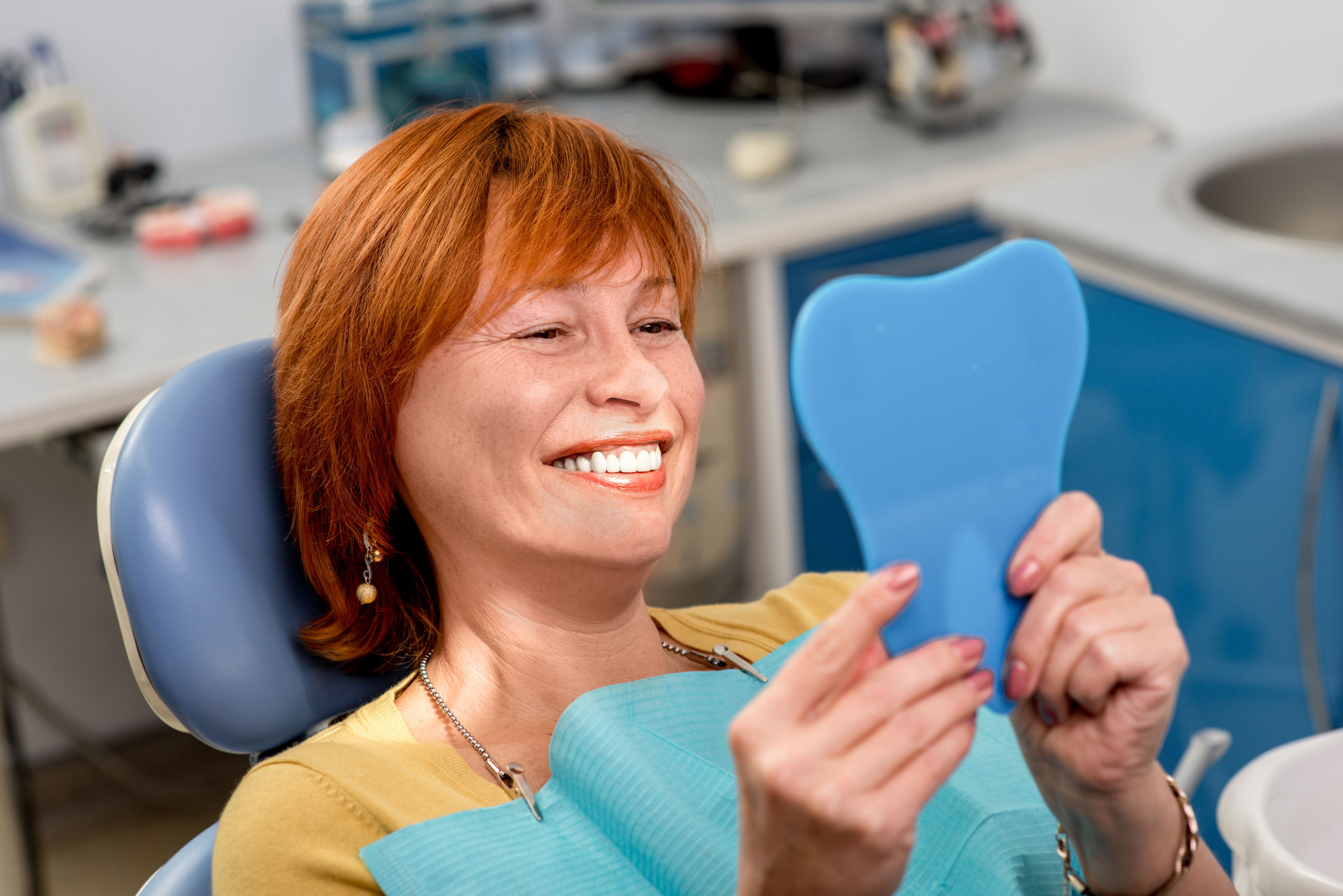 Older woman sitting in dentist's chair and smiling into handheld mirror